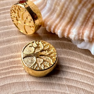 Könnte beinhalten: Zwei runde, goldfarbene Metallperlen, die jeweils ein detailliertes, geprägtes Baumdesign auf der flachen Oberfläche aufweisen. Die Perlen haben eine polierte, reflektierende Oberfläche und ein kleines Loch an der Seite, das zum Auffädeln dient.