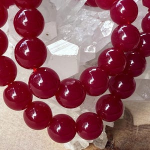 Pode incluir: Close-up de um fio de contas facetadas, vermelho escuro. As contas são redondas e enfiadas, refletindo a luz. O fundo é uma mistura de branco e castanho.