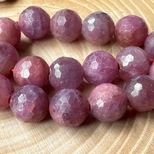 Op de afbeelding: Een streng facetgeslepen paarse edelsteen kralen. De kralen zijn rond en hebben een gladde, gepolijste afwerking. De kralen zijn geregen op een rode draad.