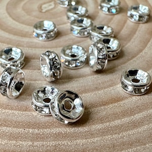 Könnte beinhalten: Nahaufnahme von silberfarbenen Metallperlen mit klaren Strasssteinen. Die Perlen sind rund und haben ein Loch in der Mitte. Sie sind in einem Kreis auf einer Holzoberfläche angeordnet.