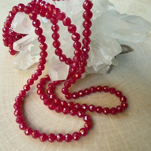 Op de afbeelding: Een lange, gefacetteerde rode kralenketting is gedrapeerd over een cluster van witte kristallen. De kralen hebben een glanzende, gepolijste afwerking. De ketting wordt gepresenteerd op een lichtgekleurd houten oppervlak.