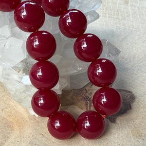 Könnte beinhalten: Eine Halskette aus großen, runden, glänzenden, tiefroten Perlen. Die Perlen sind auf einer roten Schnur aufgereiht. Der Hintergrund zeigt klare, kristalline Formationen.
