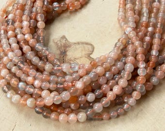 May include: A collection of smooth, round sunstone beads arranged in a circular pattern. The beads exhibit a translucent quality with varying shades of peach, soft orange, and pale cream, featuring natural internal inclusions and a subtle shimmer.