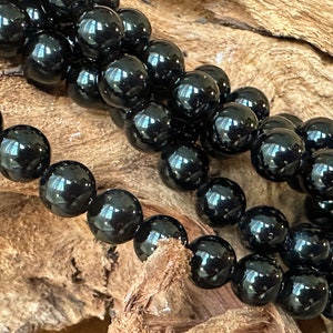 May include: A strand of smooth, spherical black beads with a polished, reflective surface. The beads are uniform in size and shape, arranged in a multi-layered coil, showcasing a deep, solid black color with bright white light reflections.