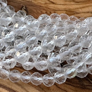 Könnte beinhalten: Eine Ansammlung von klaren, facettierten, runden Perlen, die aufgereiht sind. Jede Perle weist mehrere flache Oberflächen auf, die das Licht reflektieren und ein funkelndes Aussehen erzeugen. Die Perlen sind transparent, gleichmäßig geformt und liegen in mehreren Strängen.