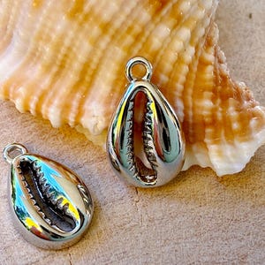 Könnte beinhalten: Zwei glänzende, silberne Kaurimuschel-Anhänger mit einer Schlaufe zum Befestigen an Schmuck. Die tropfenförmigen Anhänger haben detaillierte Innenseiten und werden auf einer hellbraunen Oberfläche mit einer Muschel präsentiert.