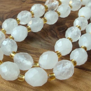May include: A necklace featuring multiple strands of faceted white translucent beads interspersed with small, cylindrical metallic gold-toned spacer beads. The beads are arranged in a repeating pattern, creating a textured appearance throughout the piece.