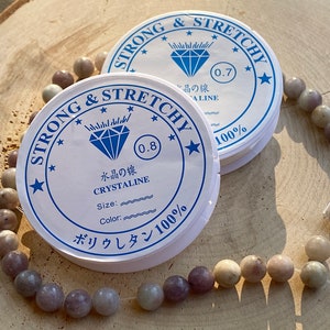 May include: Two spools of white elastic cord with blue and black text. The text on the spools reads "Strong & Stretchy", "Crystaline", "Size:", "Color:", "0.7", "0.8", "100%", and "ボリウレタン100%". A strand of light purple and gray beads is strung on the elastic cord.