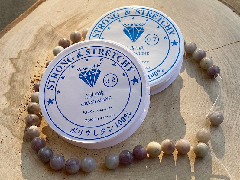 May include: Two spools of white elastic cord with blue and black text. The text on the spools reads "Strong & Stretchy", "Crystaline", "Size:", "Color:", "0.7", "0.8", "100%", and "ボリウレタン100%". A strand of light purple and gray beads is strung on the elastic cord.