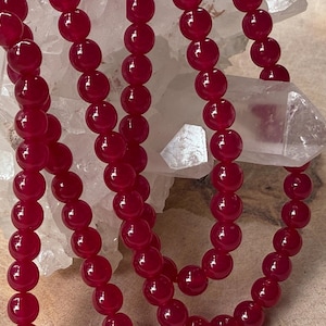 Op de afbeelding: Close-up van strengen dieprode, glanzende kralen. De kralen zijn rond en uniform van grootte, geregen om kettingen of armbanden te vormen. De achtergrond heeft een witte, kristallijne structuur, mogelijk kwarts.