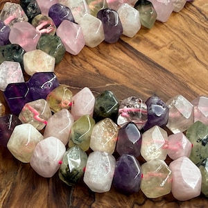 May include: A close-up of a strand of faceted gemstone beads in various colors, including purple, pink, green, and clear. The beads are strung on a pink cord, and the background is a wooden surface.