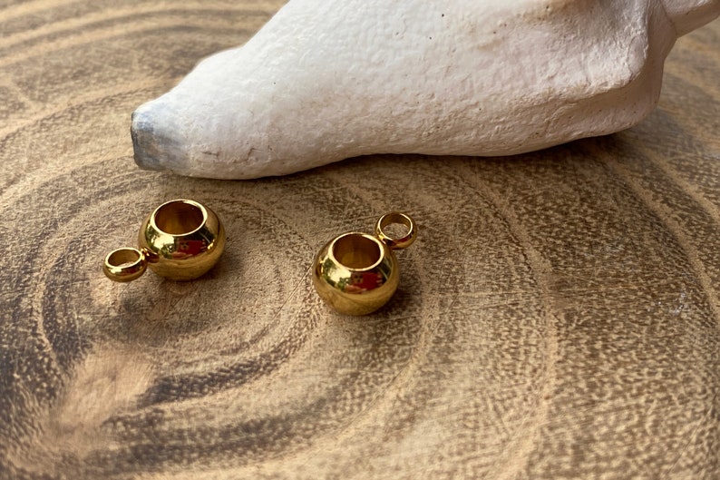 May include: Two small, spherical gold-toned metal beads, each featuring a smooth, reflective surface and a single attached jump ring loop extending from the side, designed for use as jewelry components or decorative accents in craft projects.