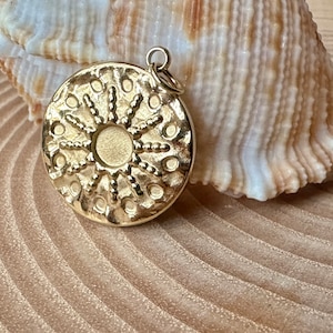 Op de afbeelding: Een goudkleurige hanger met een zonmotief. De hanger is rond met een centrale cirkel en stralende lijnen met kleine cirkels. Hij heeft een kleine lus aan de bovenkant voor een ketting. De hanger ligt op een lichtbruin oppervlak.