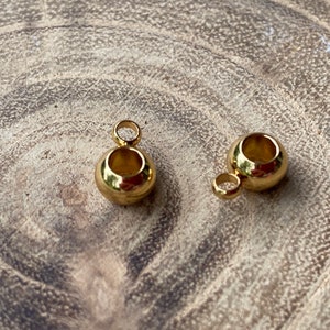 May include: Two small, spherical gold-toned metal beads, each featuring a smooth, polished surface and a central hollow opening. Each bead is attached to a small, circular jump ring loop at the top, designed for use as a jewelry pendant or charm.