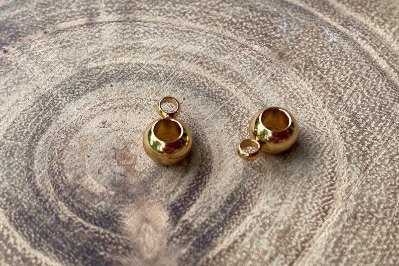 May include: Two small, spherical gold-toned metal beads, each featuring a smooth, polished surface and a central hollow opening. Each bead is attached to a small, circular jump ring loop at the top, designed for use as a jewelry pendant or charm.