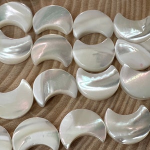 Op de afbeelding: Een verzameling witte, iriserende parelmoer halve maanvormige kralen. De kralen zijn glad en gepolijst met een subtiele glans.