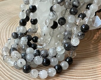 May include: A collection of round beads strung together, featuring a mix of clear, translucent, and opaque black stones. Many of the clear beads contain visible black needle-like inclusions, creating a textured, high-contrast appearance.