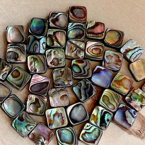 Könnte beinhalten: Eine Sammlung quadratischer Abalone-Muschelkernperlen. Die Perlen zeigen schillernde Farben von Grün, Blau, Lila und Braun. Die Abalone-Muscheln haben ein natürliches, wirbelndes Muster und sind auf einer hellbraunen Oberfläche angeordnet.
