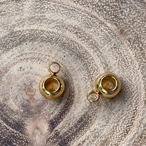May include: Two small, polished gold-tone metal beads, each featuring a smooth, spherical shape with a central hole and a small, circular jump ring attached to the top, designed for use as jewelry components or decorative charms.