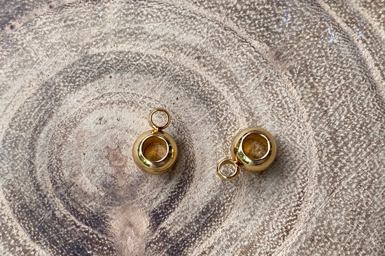 May include: Two small, polished gold-tone metal beads, each featuring a smooth, spherical shape with a central hole and a small, circular jump ring attached to the top, designed for use as jewelry components or decorative charms.