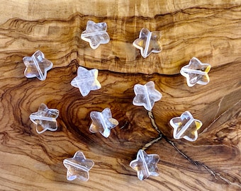 May include: Clear, star-shaped crystal beads are scattered on a wooden surface. The beads have a translucent appearance with visible internal structures. The wooden surface has a natural, warm brown tone with visible grain patterns. The stars are approximately 2.5 cm in diameter.