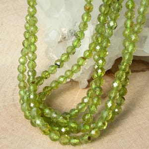 May include: A strand of faceted, translucent lime green peridot beads. The small, round, polished gemstones are strung together in a continuous loop, showcasing a vibrant, consistent color and a reflective, multi-faceted surface texture.