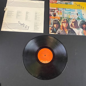 May include: An album cover and vinyl record by the band Sweet. The album cover features the band members with a city scene background. The vinyl record is black with an orange label. The album's title is visible on the cover.