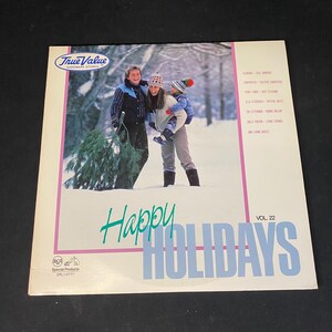 May include: A vintage vinyl record album cover with the text "Happy Holidays" in teal and blue. The cover features a winter scene with three people carrying a Christmas tree. The album is labeled "Vol. 22" and includes a list of artists.