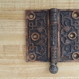 May include: Antique brass door hinge with ornate floral design. The hinge is closed and has a round knob on the bottom.