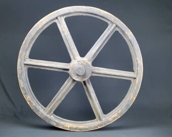 May include: A large, wooden wagon wheel with six spokes. The wheel is painted a dark gray color and has a natural wood finish on the rim.