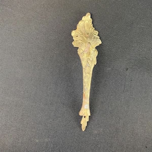 May include: A vintage, ornate brass furniture pull with a floral design. The pull has a detailed, textured surface and a long, slender handle. The overall color is a warm, antique gold, with some areas showing signs of age and wear.