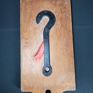 May include: A wooden board with a black metal hook and a red wooden handle attached to it. The hook and handle are mounted on the board.