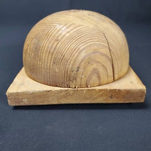 May include: A wooden hat block with a round top and a square base. The block is made of light brown wood with a visible grain pattern.