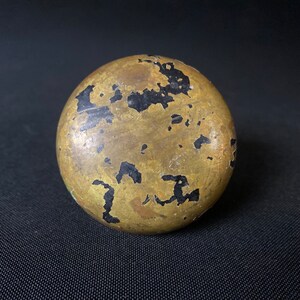 May include: A round, brass-colored object with black paint splatter. The object has a smooth, slightly textured surface.