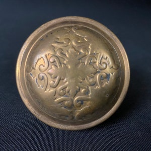 May include: A round, antique brass knob with an ornate, raised floral design in the center. The knob has a slightly domed shape and a detailed, textured surface. The edges show signs of wear, adding to its vintage appeal.