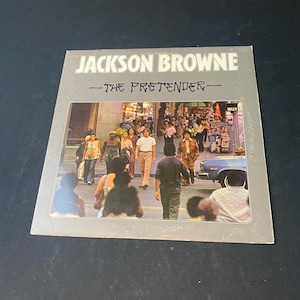 May include: Album cover for "The Pretender" by Jackson Browne. The cover features a photograph of a crowd of people walking on a city street. The album title and artist name are in a gray box at the top.