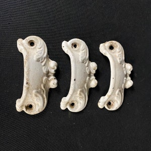 May include: Three white ceramic drawer pulls with a decorative, ornate design. Each pull has a curved shape with a raised, floral pattern.