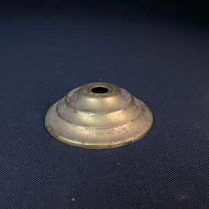 May include: A gold-toned metal lamp finial with a tiered, stepped design. The finial has a small hole in the center.