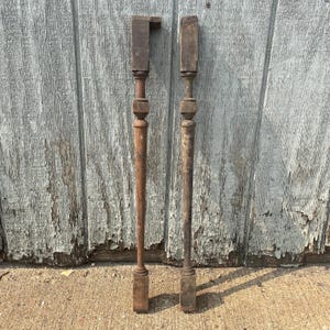 May include: Two wooden balusters with a square top and a decorative design. The balusters are brown and have a distressed finish.