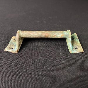 May include: A weathered bronze-colored handle with a rectangular bar and two mounting plates. The handle shows signs of age with a greenish patina, and each plate has two screw holes. The hardware is likely for a cabinet or drawer.