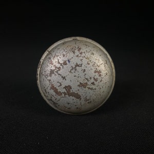 May include: A silver-colored, round, metal knob with a worn, textured surface. The knob has a slightly raised edge.