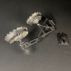 May include: Black cast iron wall mount with two ornate circular brackets and a decorative plate. The brackets have a detailed design with a central knob and a scalloped edge.