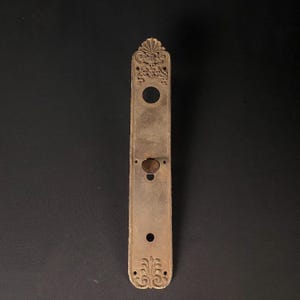May include: An antique, ornate, metal door plate with a keyhole and a decorative design at the top and bottom.