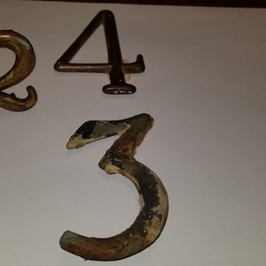 May include: Three metal house numbers, 2, 3, and 4, in a vintage style. The numbers are made of brass and have a worn, aged look. The number 3 is the most worn and has a green patina.