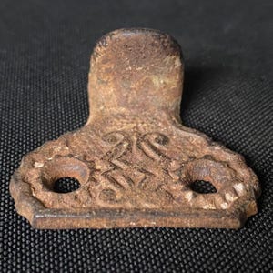 May include: A rusty metal door hinge with two holes and an ornate design. The hinge is a dark brown color and has a slightly curved shape.