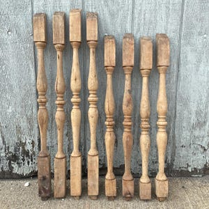 May include: Eight wooden balusters, each with a unique turned design, are arranged in a row against a gray background. The balusters are all approximately the same length and width, and they have a natural wood finish.