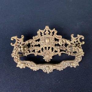 May include: Ornate brass drawer pull with a decorative, intricate design. The pull has a curved shape and a central, raised section.