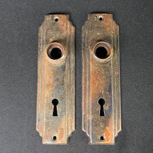 May include: Two antique, rusty, metal door plates with a keyhole and a round hole in the center. The plates have a decorative, art deco design.
