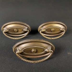 May include: Three antique brass oval cabinet door pulls with ornate detailing. Each pull has a raised oval center with a small round knob.