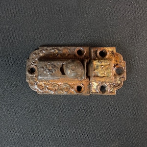 May include: A rusty metal latch with an ornate design and a round knob. The latch is made of cast iron and has a dark brown color.